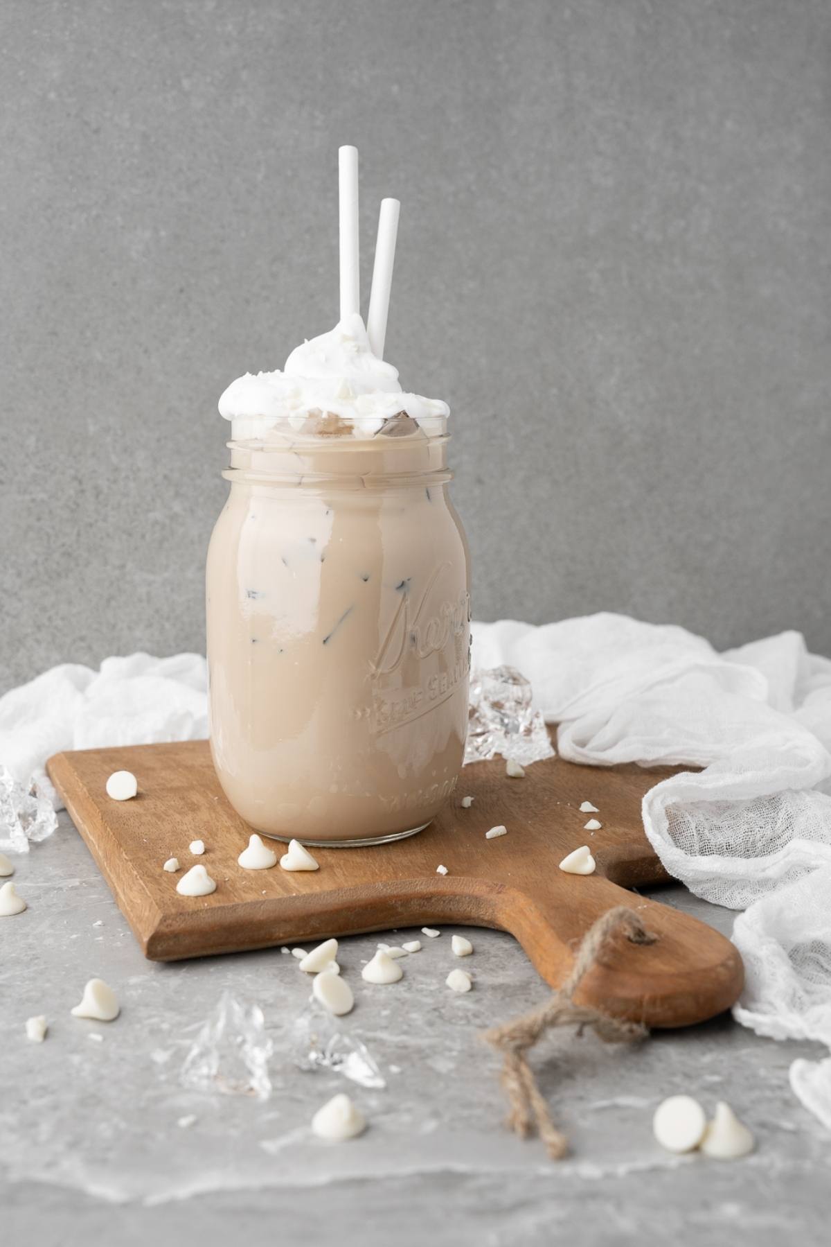 Iced coffee with whipped cream and two straws in a mason jar on a wooden board, with white chocolate chips—perfect for your DIY white chocolate mocha iced coffee craving.