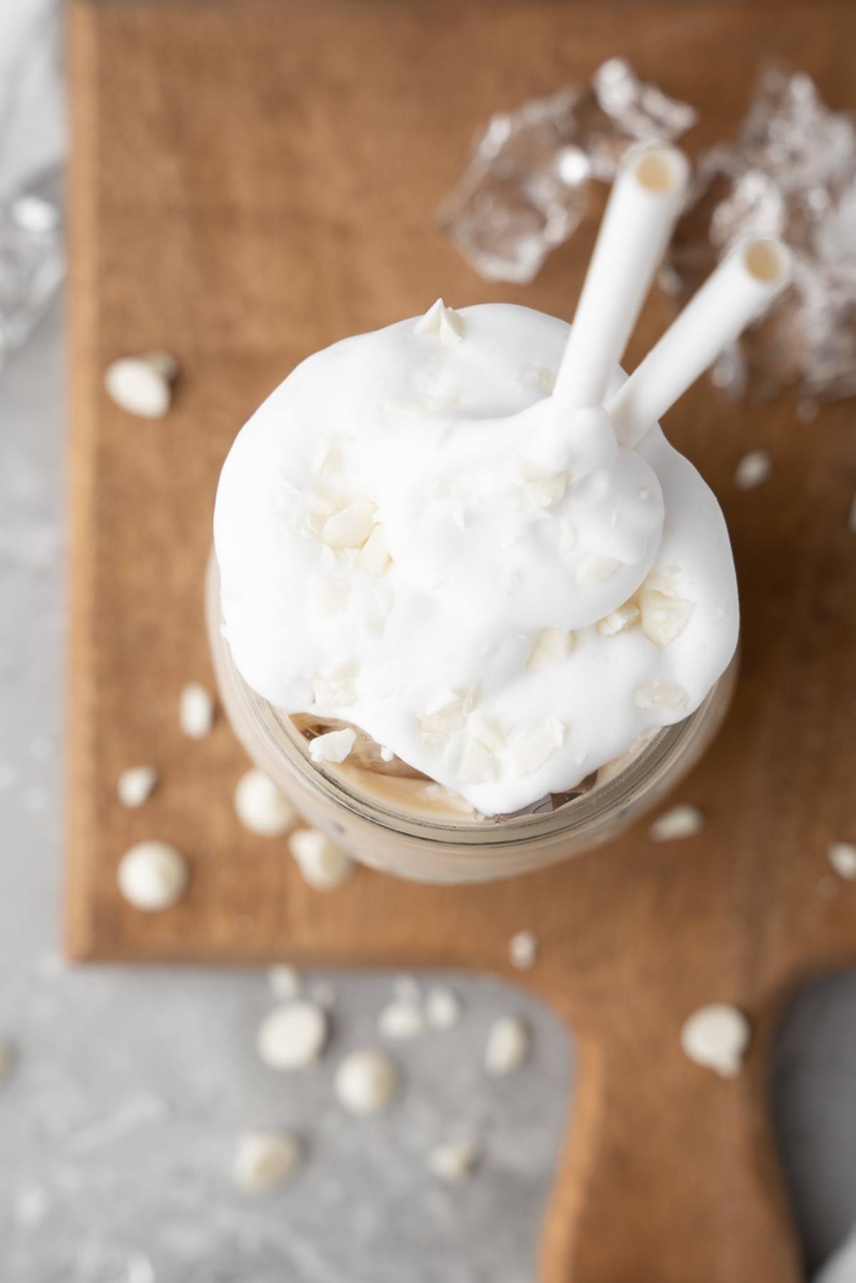 Mason jar filled with whipped cream, white chocolate chips, and two straws on a wooden board—perfect for your DIY white chocolate mocha iced coffee or Starbucks Copycat Iced White Chocolate Mocha at home.