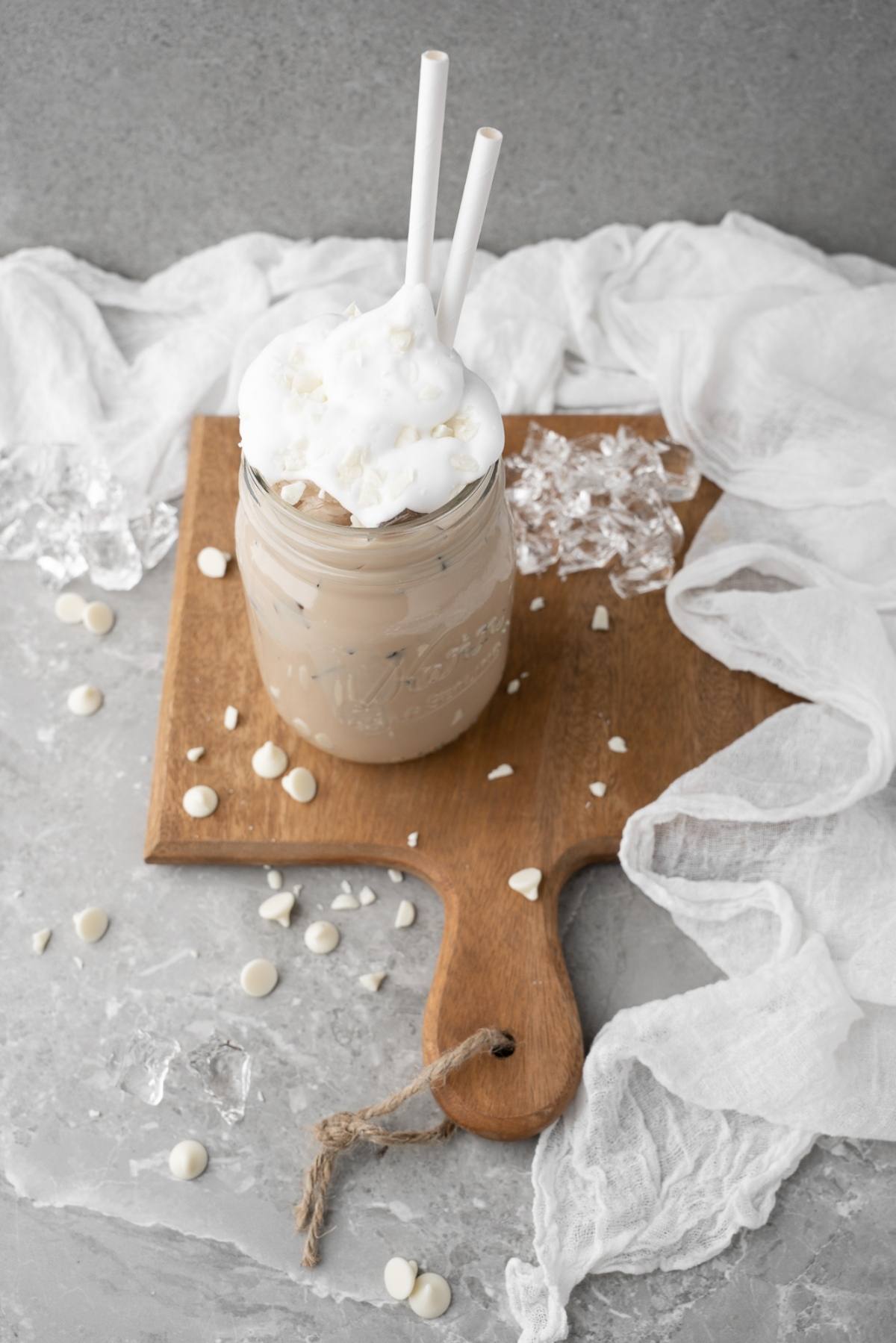 Homemade iced mocha topped with whipped cream in a jar, two straws, served on a wooden board with ice and white chocolate chips—your DIY white chocolate mocha iced coffee at home.