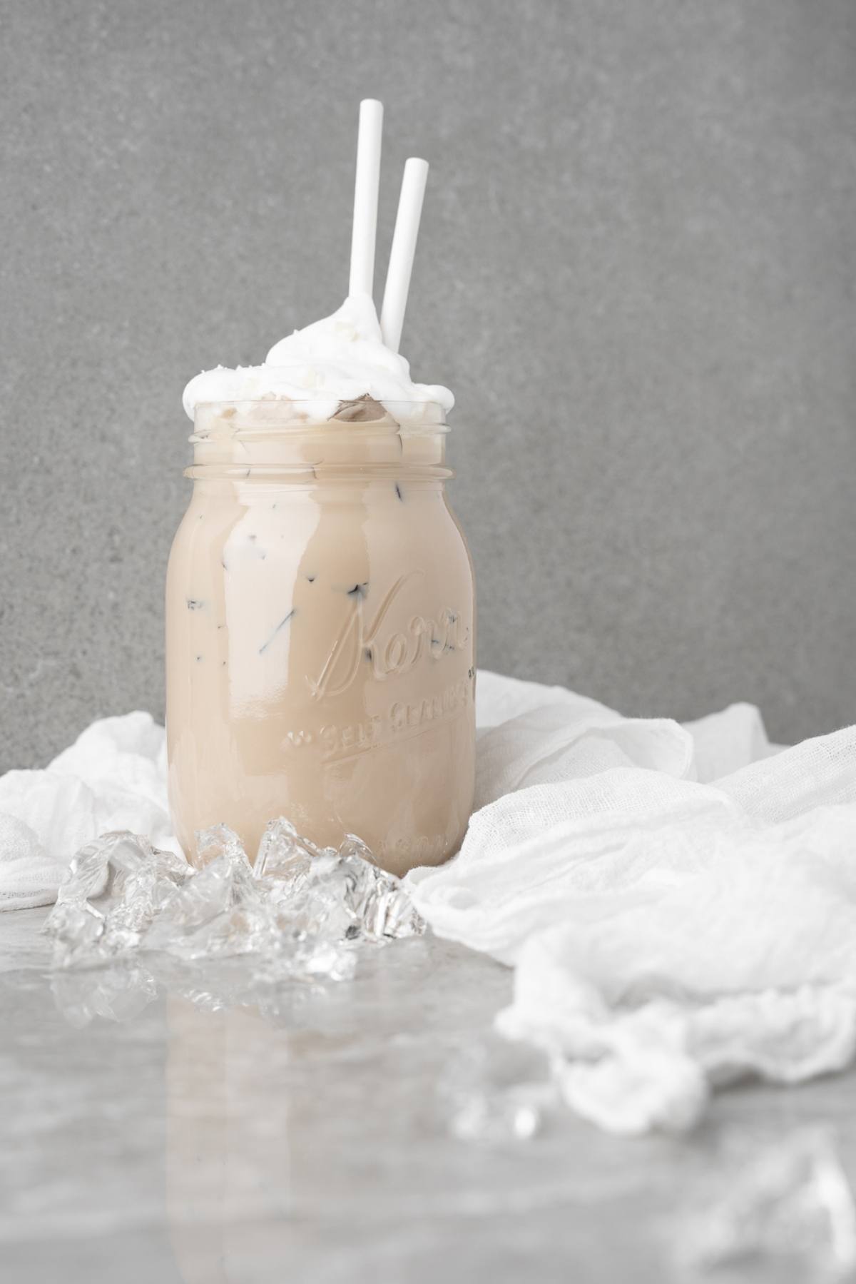 Iced coffee with whipped cream in a mason jar, two straws, and ice cubes on a marble surface—a perfect DIY white chocolate mocha iced coffee inspired by your favorite café treat.