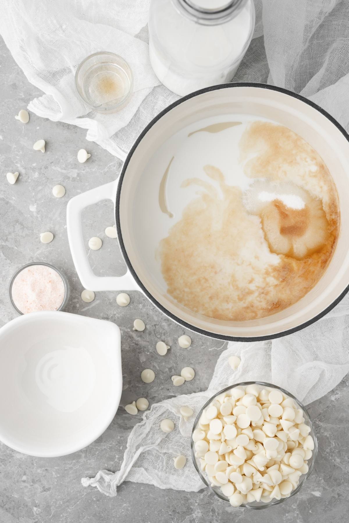 White chocolate chips, milk, sugar, and vanilla in and around a pot on a gray countertop—perfect for making a DIY white chocolate mocha iced coffee at home.