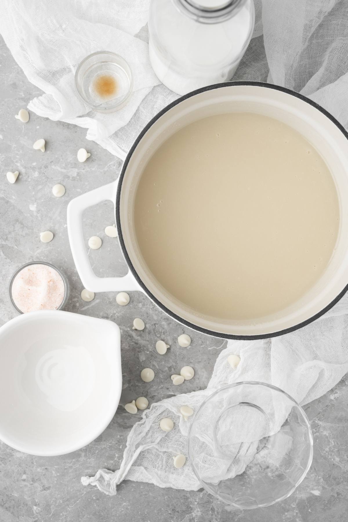 A pot of white liquid surrounded by white chocolate chips, a pitcher, glass, and small bowls on a gray surface—perfect for making a DIY white chocolate mocha iced coffee at home.