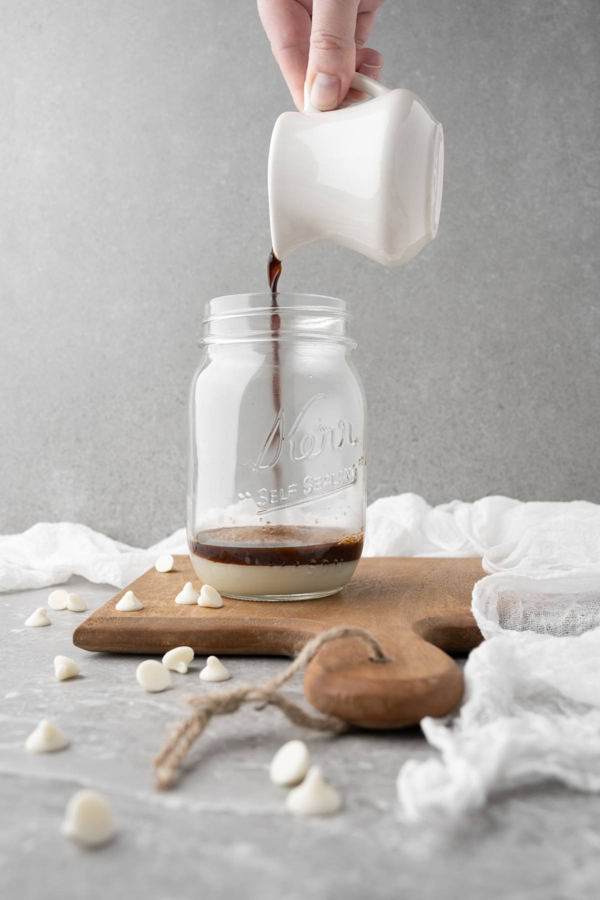 Hand pours dark liquid from a small pitcher into a mason jar with white chips on a wooden board, creating a DIY white chocolate mocha iced coffee.