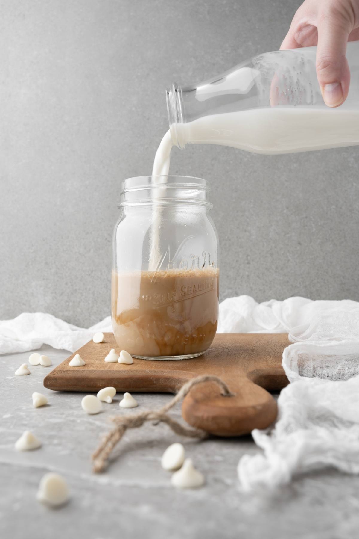 A hand pours milk from a bottle into a mason jar with coffee, white chocolate chips scattered around—a perfect homemade iced mocha for those who love enjoying Starbucks iced coffee at home.