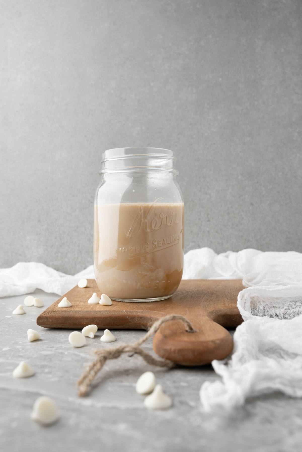 A mason jar of creamy liquid sits on a wooden board, surrounded by white chocolate chips—perfect for making a homemade iced mocha or your own Starbucks Copycat Iced White Chocolate Mocha at home.