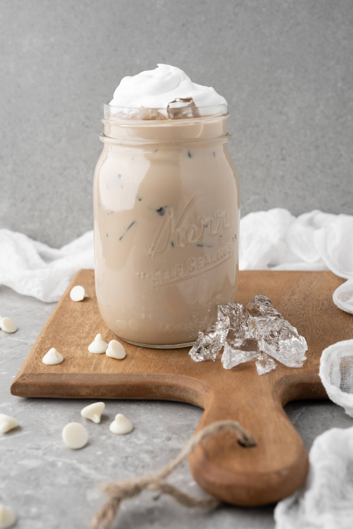 Enjoy a DIY white chocolate mocha iced coffee with whipped cream in a mason jar, served on a wooden board with ice and white chocolate chips—for a Starbucks iced coffee at home experience.