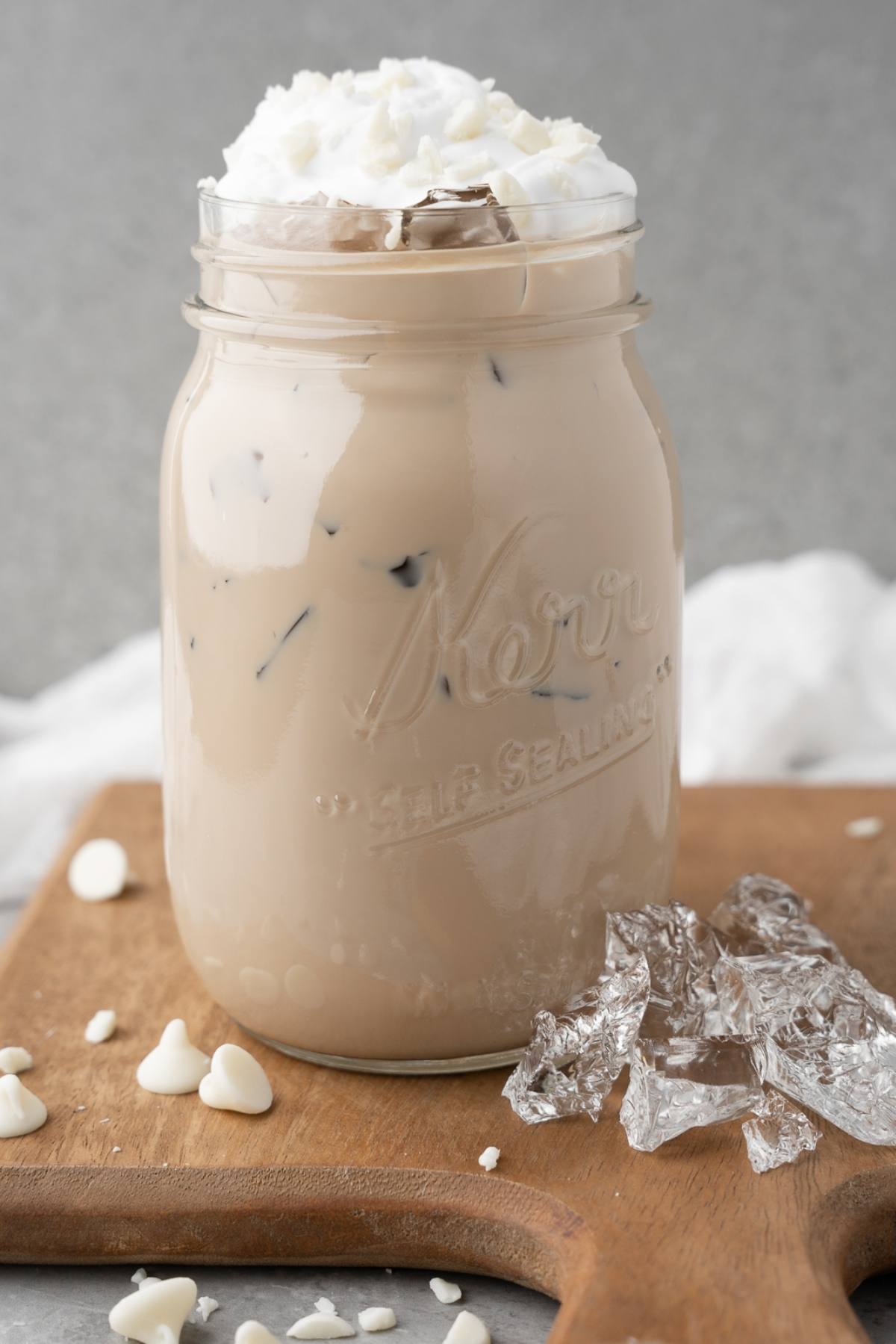Iced coffee with whipped cream in a mason jar, on a wooden board with ice and white chocolate chips—perfect for enjoying your own DIY white chocolate mocha iced coffee at home.