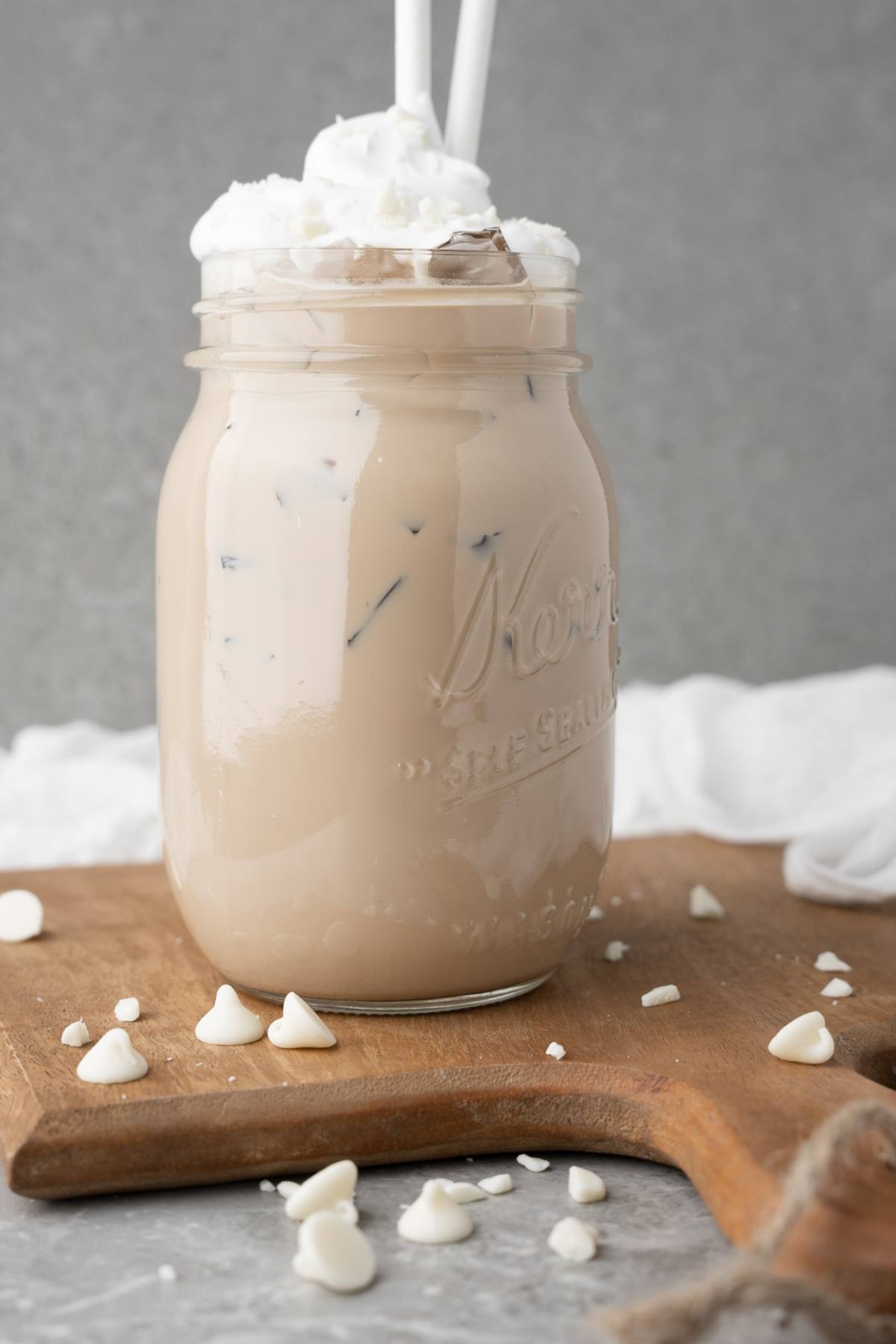 Enjoy a DIY white chocolate mocha iced coffee topped with whipped cream in a mason jar, paired with straws and white chocolate chips—your perfect copycat Iced White Chocolate Mocha to recreate Starbucks iced coffee at home.