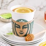 A Starbucks cup with a lemon slice on top, honey dipper, and lemon slices on a stack of white plates—perfect for making your own Starbucks Copycat Medicine Ball at home.