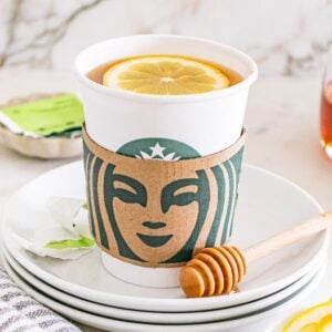 A Starbucks cup with a lemon slice on top, honey dipper, and lemon slices on a stack of white plates—perfect for making your own Starbucks Copycat Medicine Ball at home.
