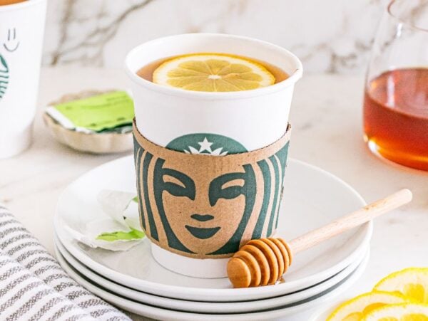 A Starbucks cup with a lemon slice on top, honey dipper, and lemon slices on a stack of white plates—perfect for making your own Starbucks Copycat Medicine Ball at home.