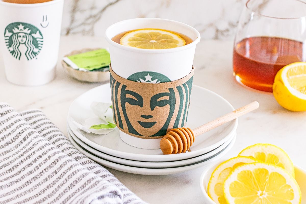Starbucks Copycat Medicine Ball with a Starbucks cup, lemon slices, honey dipper, tea bags, and lemon slices on white plates.