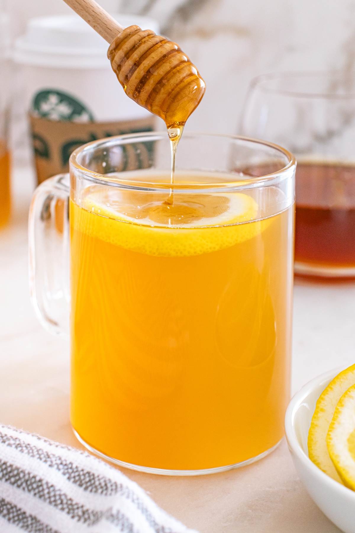 A honey dipper drizzles honey into a glass mug of tea with a lemon slice.