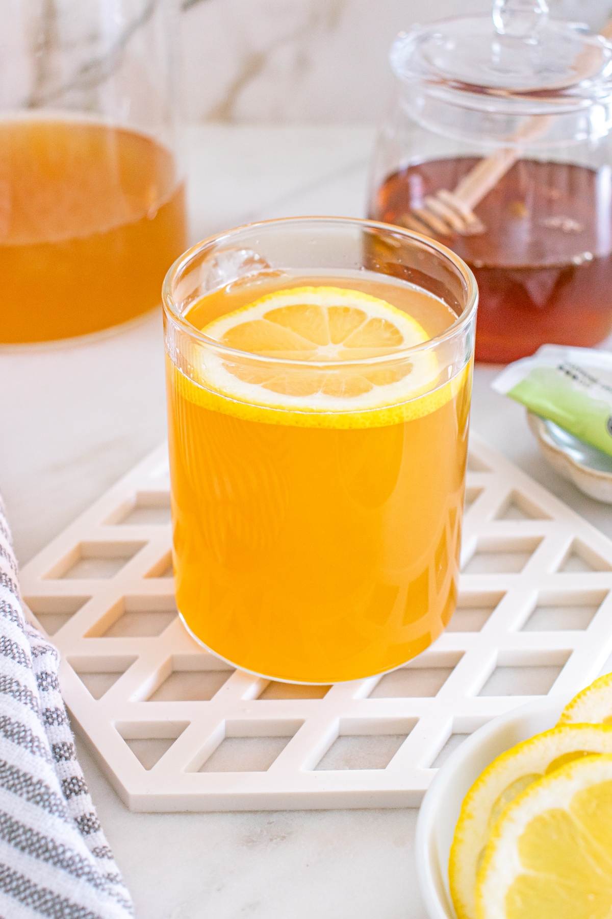 A glass of orange tea with a lemon slice, beside a honey jar and extra lemon slices on a white table.