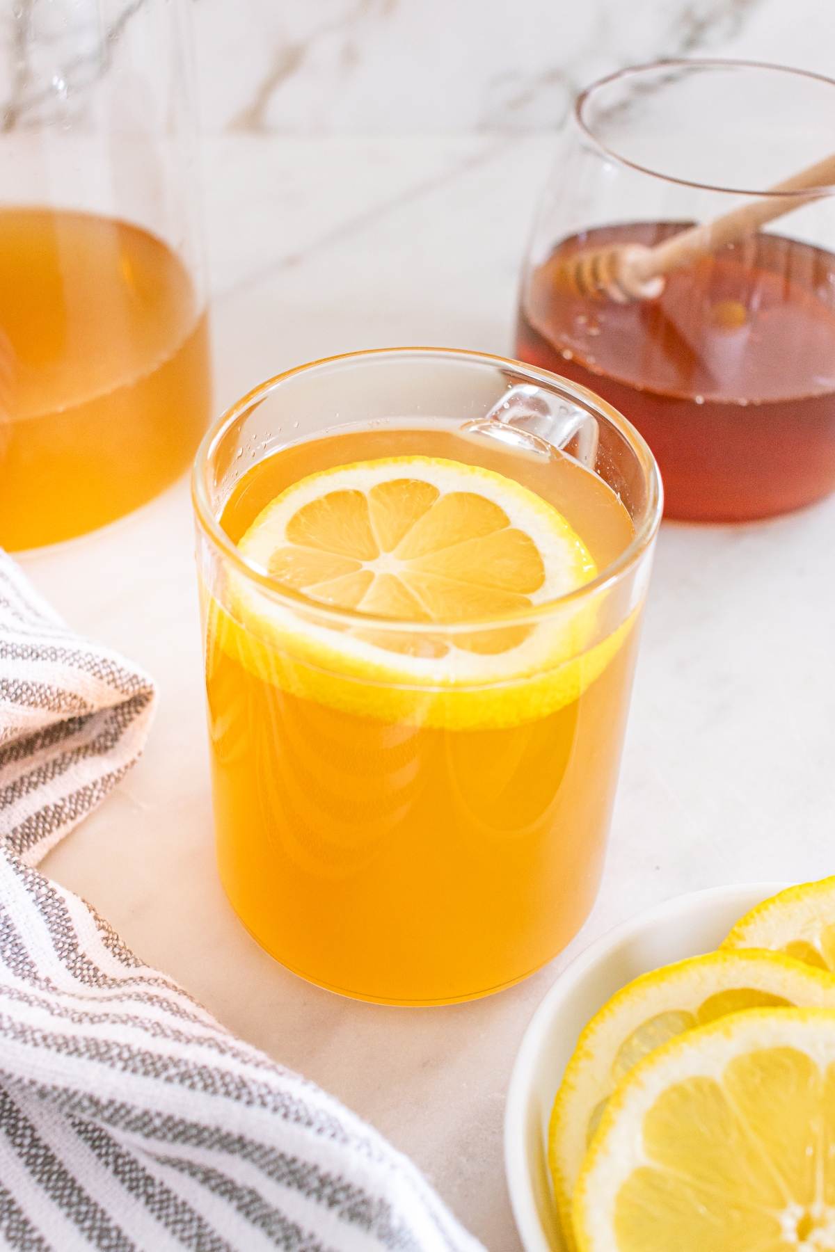 A glass mug of hot lemon tea with a lemon slice, next to honey, a pitcher, and sliced lemons on a table.