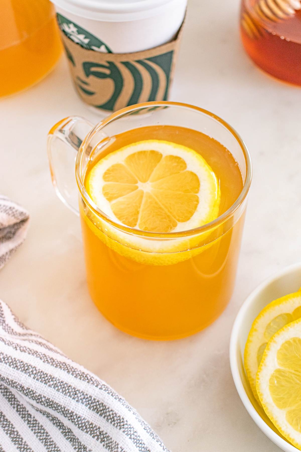 A glass mug of hot lemon tea with a lemon slice on top, next to a Starbucks cup and lemon slices.