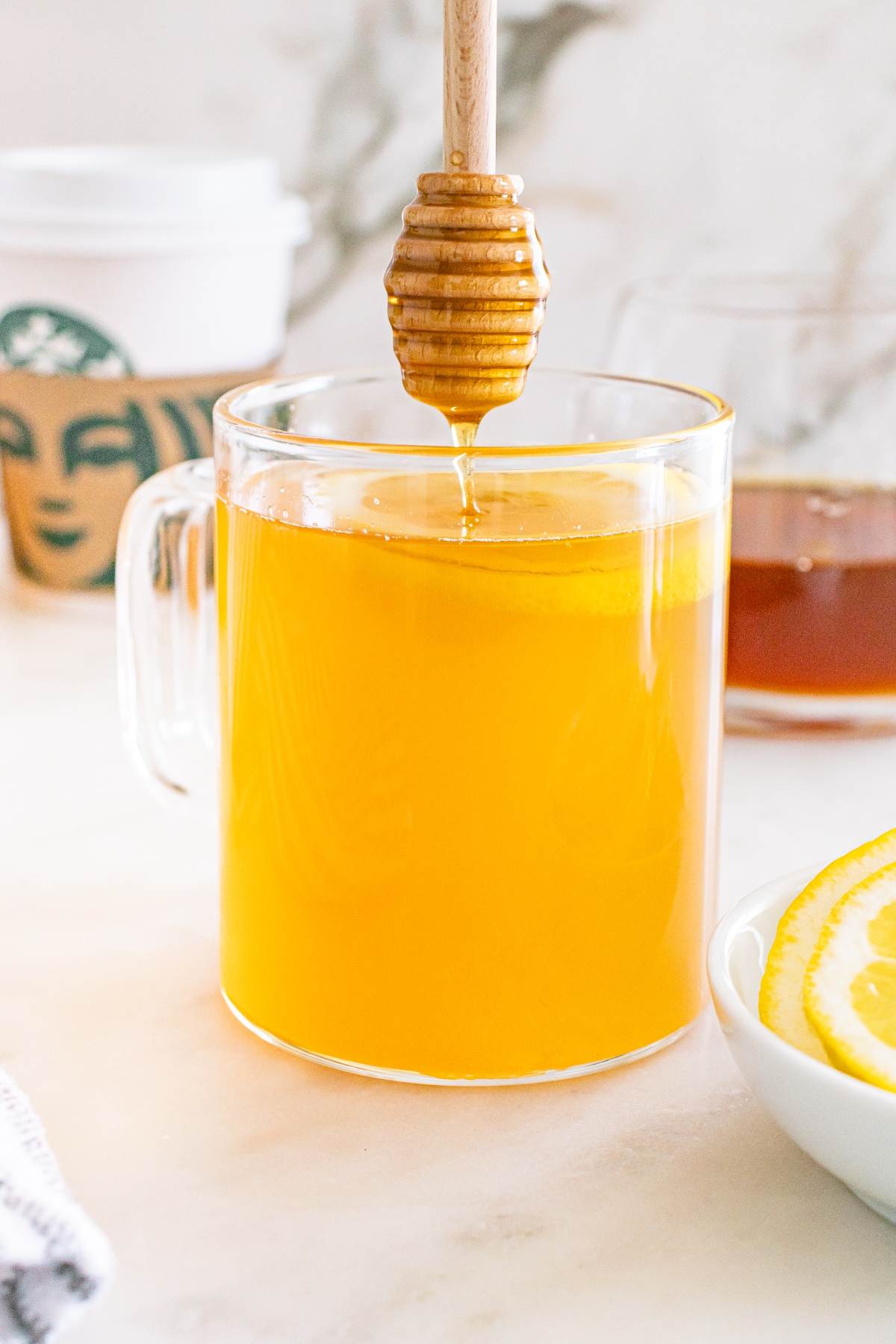 Glass mug of hot tea with a lemon slice, honey dipper above, and Starbucks cup in the background.