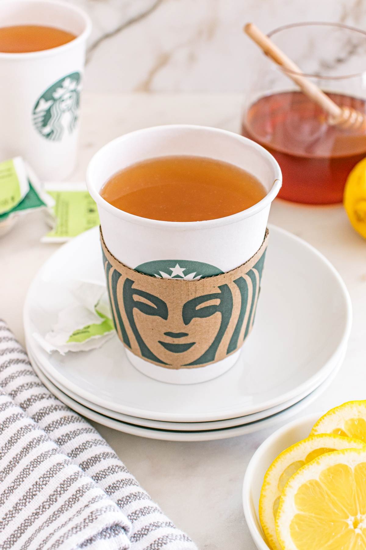 A cup of tea in a Starbucks cup with lemon slices and honey on the side.