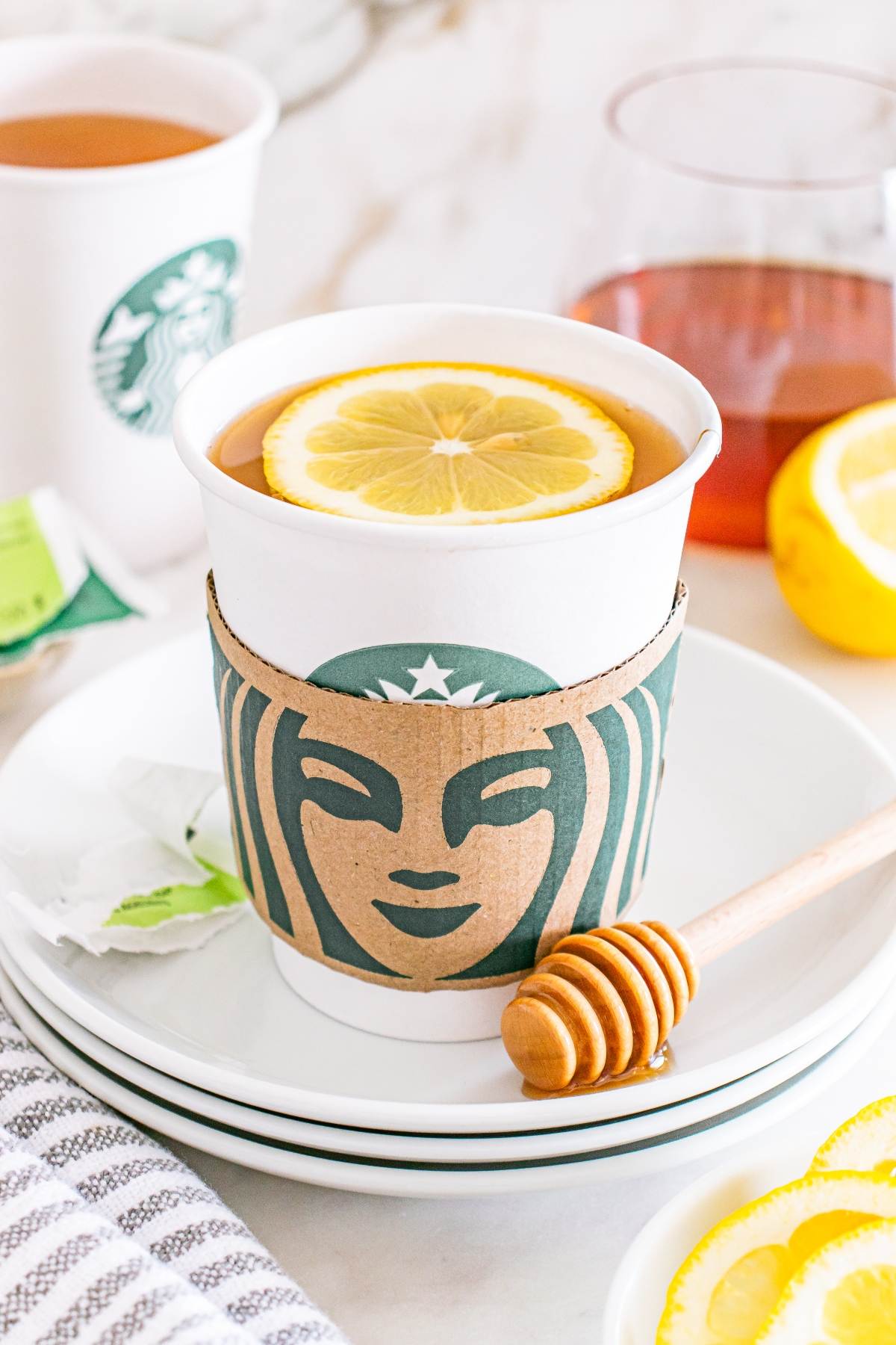 A Starbucks cup with tea, topped with a lemon slice, sits on a plate with a honey dipper and lemon slices.