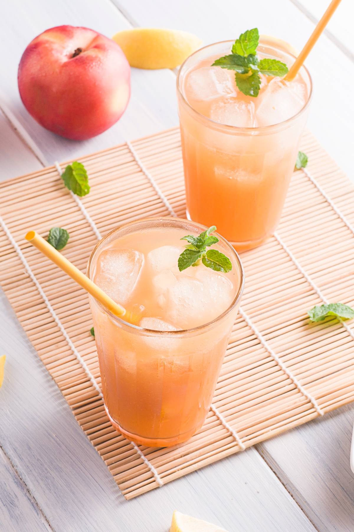 Two glasses of homemade Starbucks peach tea with mint and straws on a bamboo mat, accompanied by a peach and lemon wedges—perfect for trying your own summer Starbucks drink recipe at home.