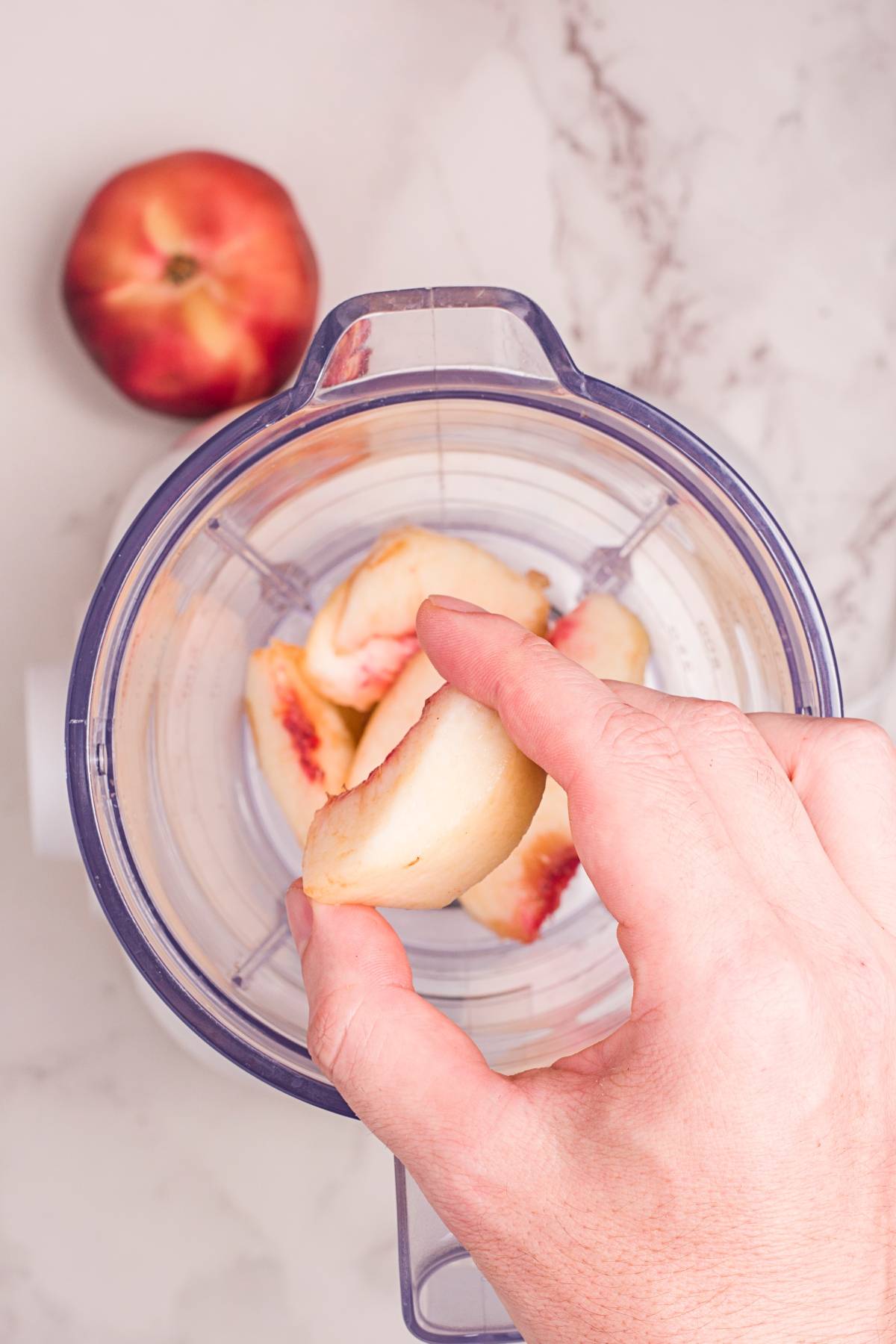 Hand holding a peach slice over a blender with more peach slices inside, whole peach on countertop—perfect for making your own homemade Starbucks peach tea or refreshing iced peach tea.