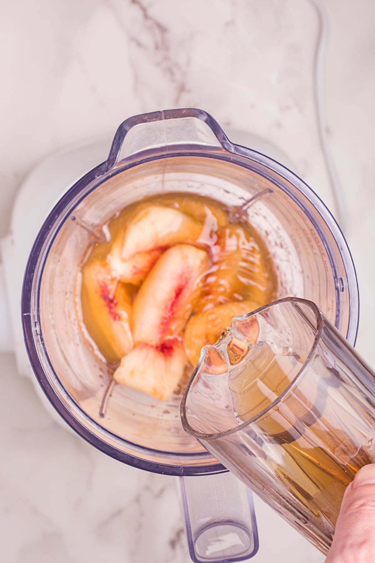 Hand pouring liquid into a blender with peach slices and juice on a marble surface, prepping a refreshing Homemade Starbucks peach tea—perfect for those craving a delicious summer Starbucks drink recipe.