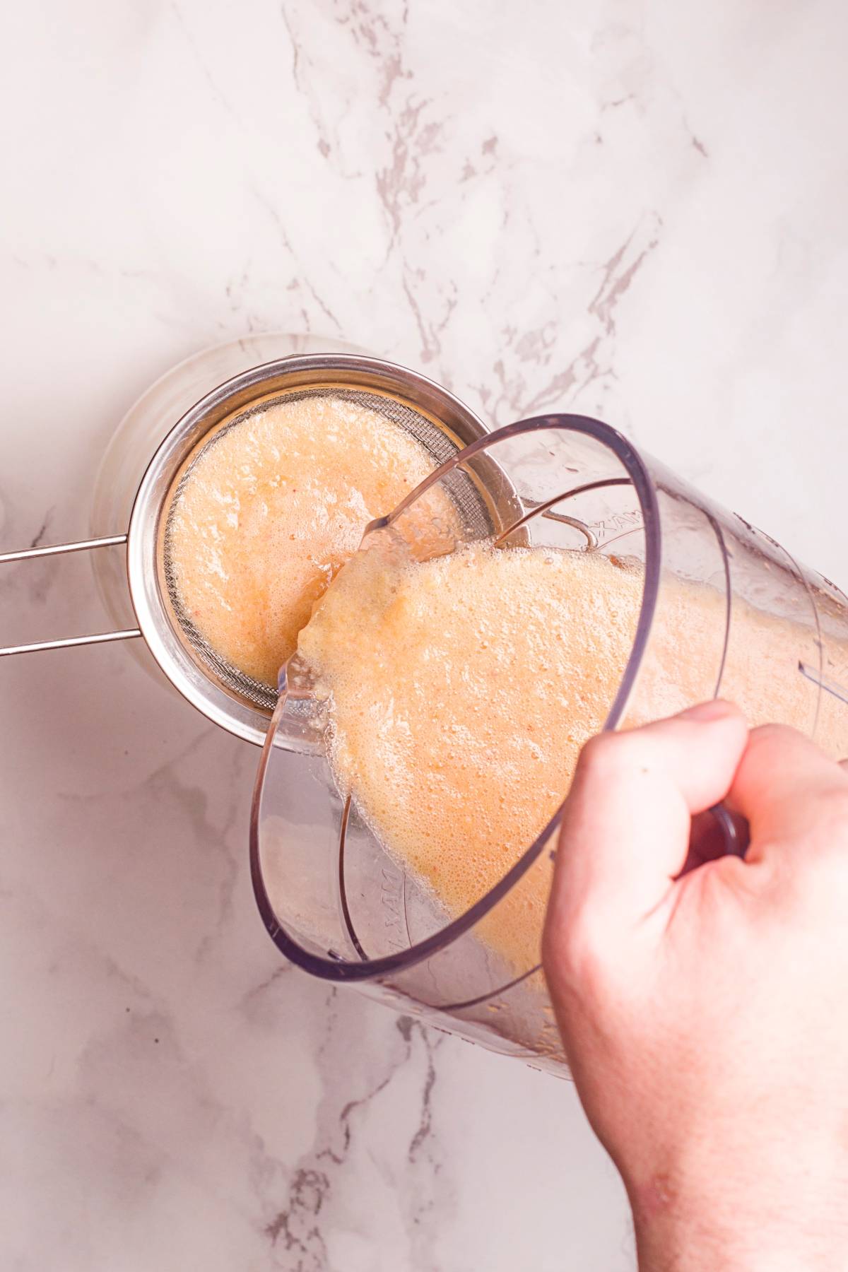 Person pouring blended fruit juice through a fine mesh strainer over a glass bowl on a marble countertop, perfect for creating your own DIY Starbucks lemonade recipe or a refreshing iced peach tea at home.