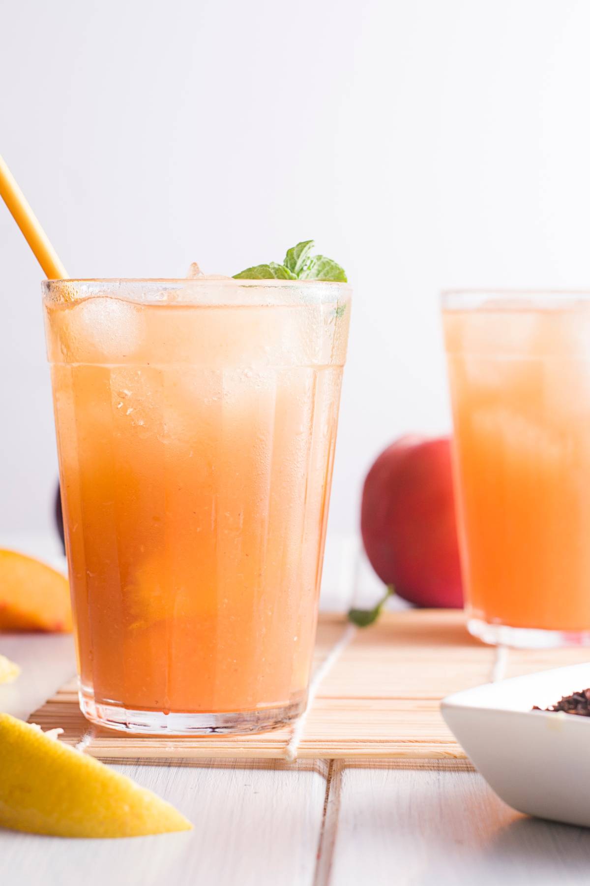 A glass of iced homemade Starbucks peach tea with a yellow straw and mint garnish, served with fresh peach slices nearby.