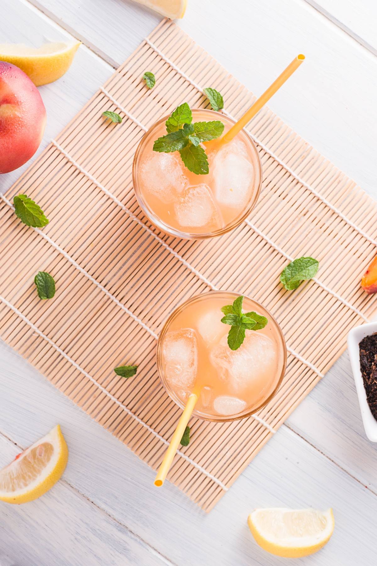 Two iced peach drinks with mint and straws on a bamboo mat, surrounded by lemon wedges and peach slices—perfect for enjoying homemade Starbucks peach tea or trying your own iced peach tea twist at home.