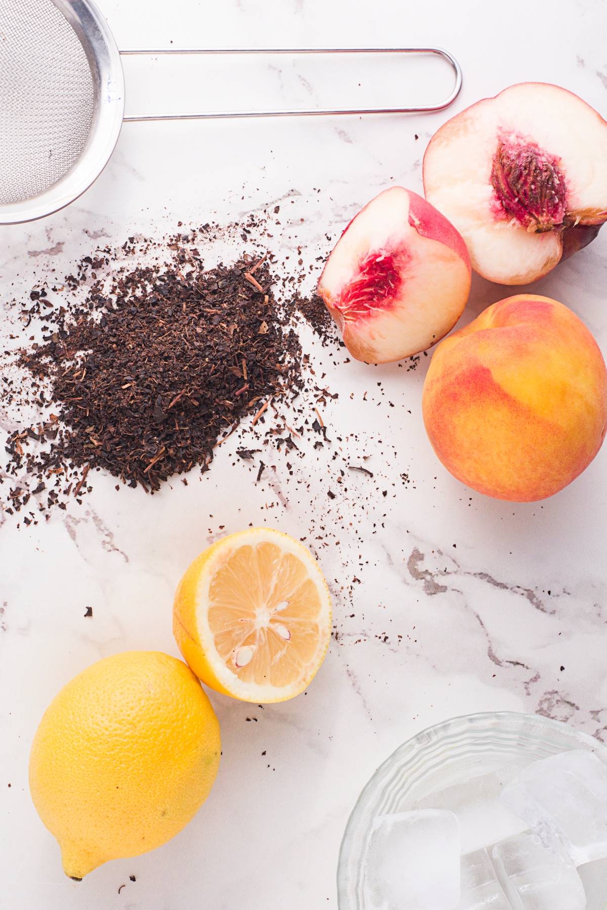 Loose tea, halved and whole peaches, lemon, a strainer, and ice water on a white surface—perfect ingredients for a refreshing Starbucks copycat peach black tea lemonade or your own iced peach tea summer Starbucks drink recipe.
