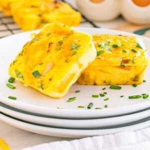 Two square Starbucks copycat potato cheddar and chive bakes garnished with chopped chives on stacked white plates, with more muffins in the background.