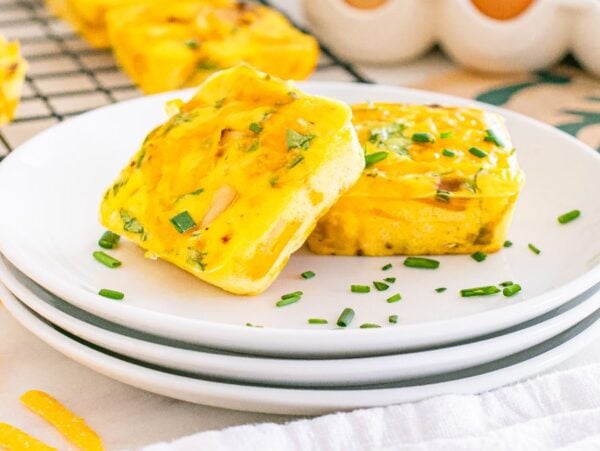 Two square Starbucks copycat potato cheddar and chive bakes garnished with chopped chives on stacked white plates, with more muffins in the background.