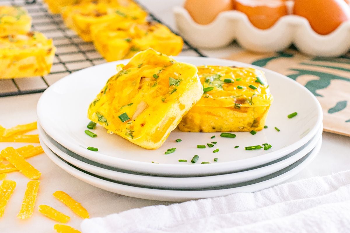 Two baked egg bites with herbs on a white plate, inspired by Starbucks copycat potato cheddar and chive bakes, with eggs and more muffins in the background.
