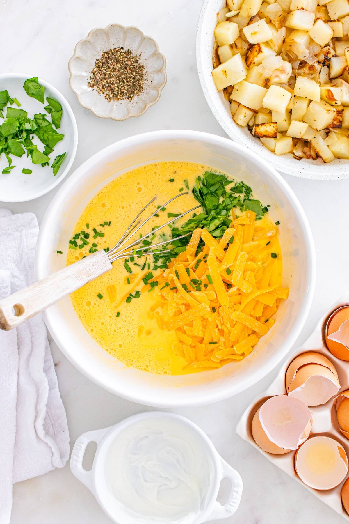 Bowl of beaten eggs with chives and cheese, surrounded by potatoes, herbs, eggshells, and pepper.
