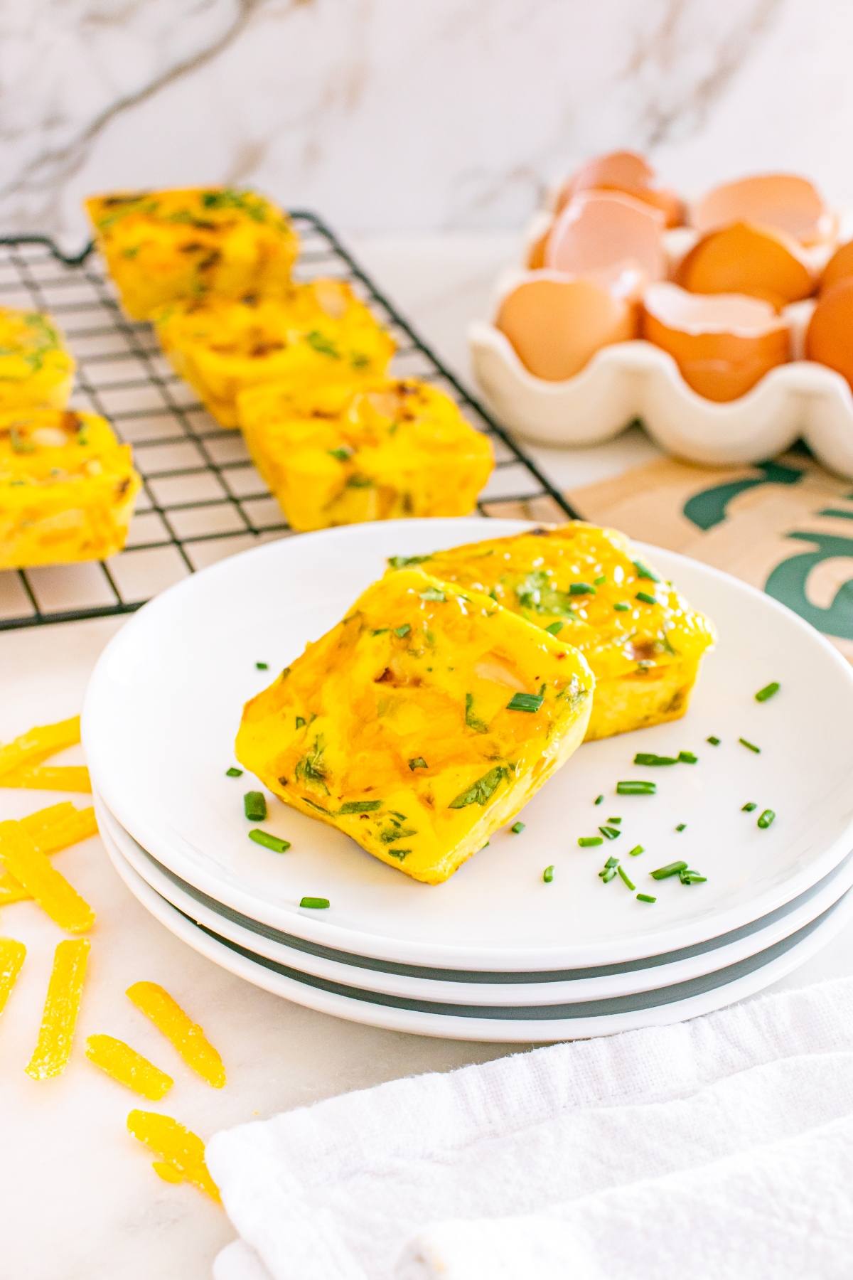 Two square egg bites with herbs on a white plate, eggshells and a cooling rack with more egg bites in the background.