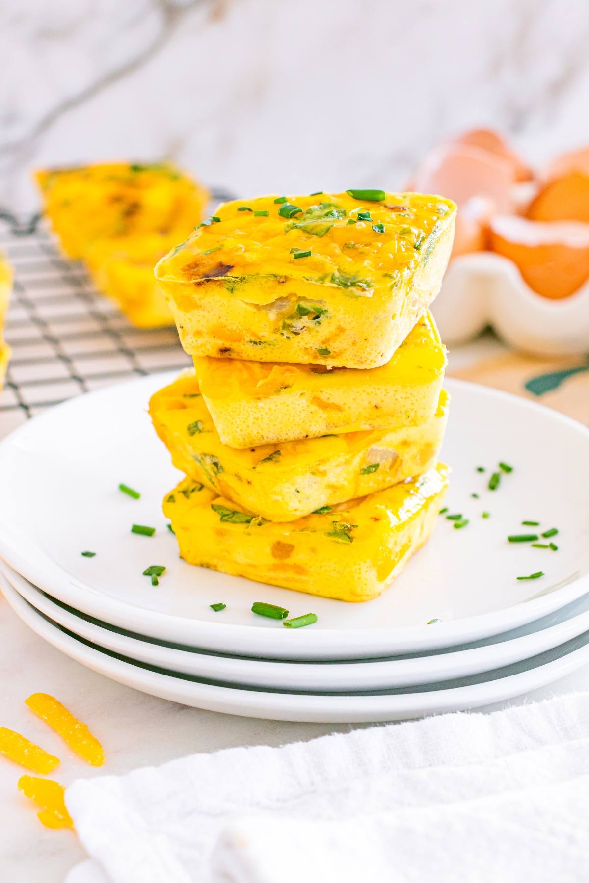 Three baked egg bites stacked on white plates, garnished with chopped chives, with eggs and more nearby.
