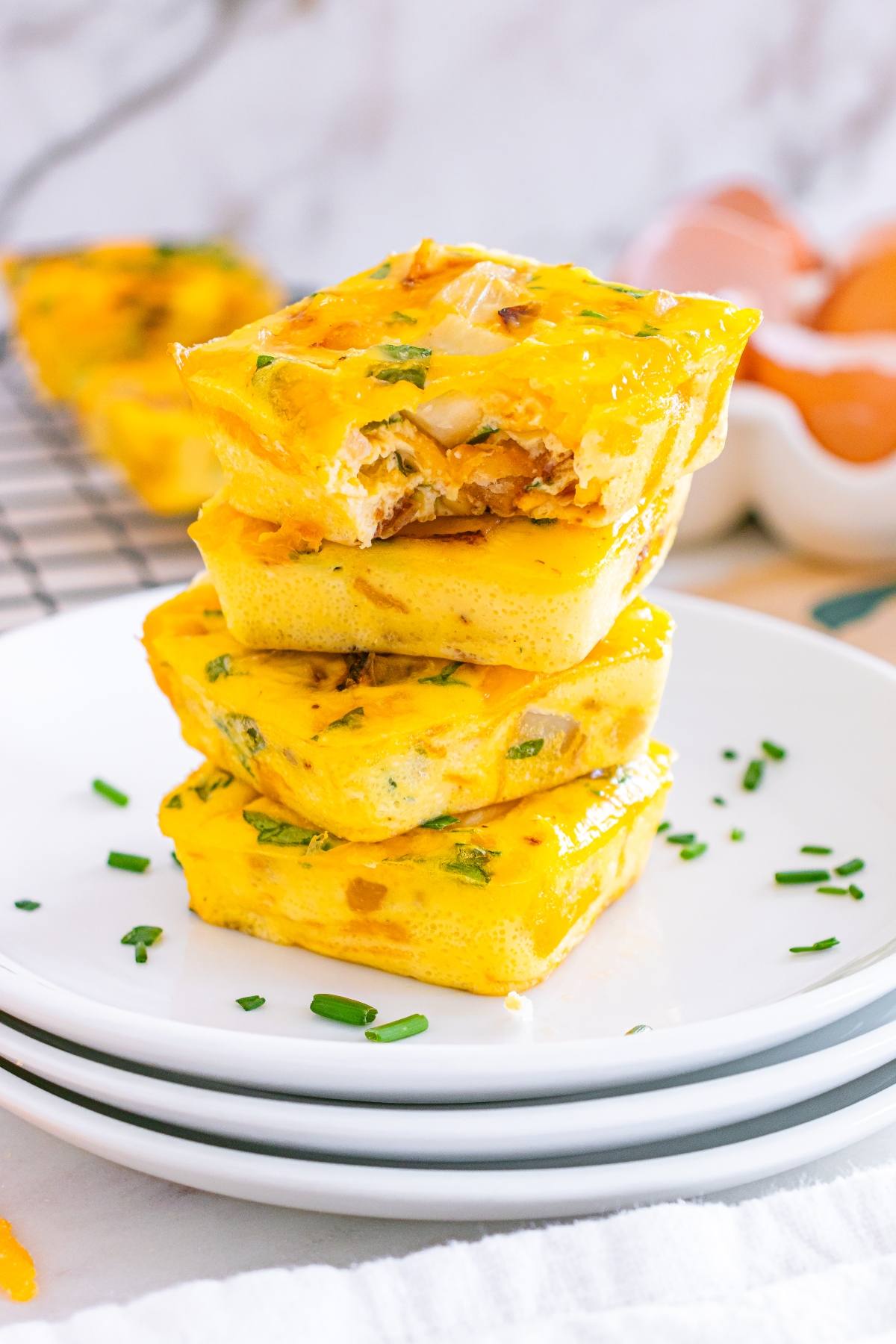 Three square egg bites stacked on white plates, garnished with chopped chives, with eggs in the background.
