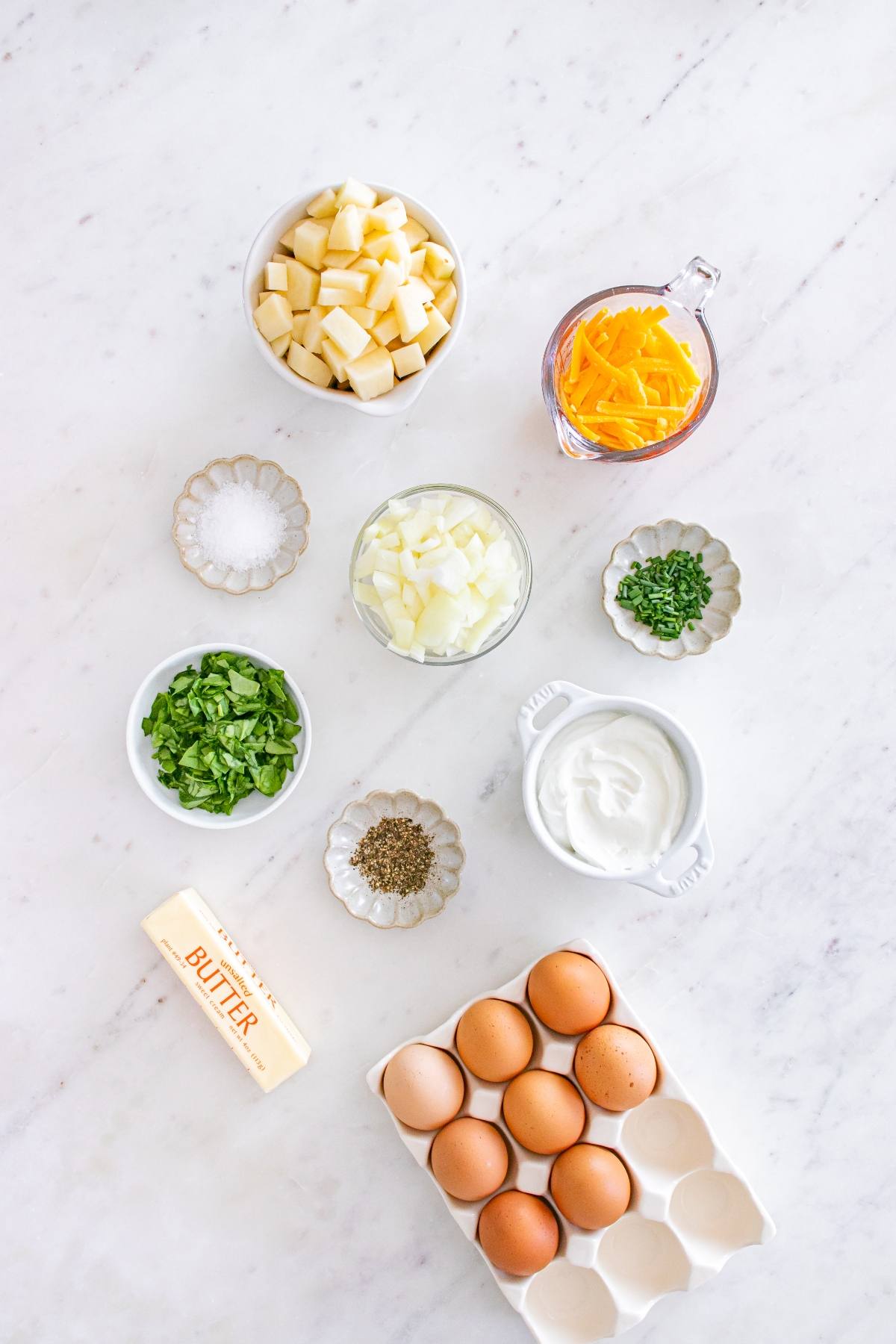Ingredients on a white surface: eggs, butter, potatoes, cheese, onion, spinach, herbs, salt, pepper, and sour cream.