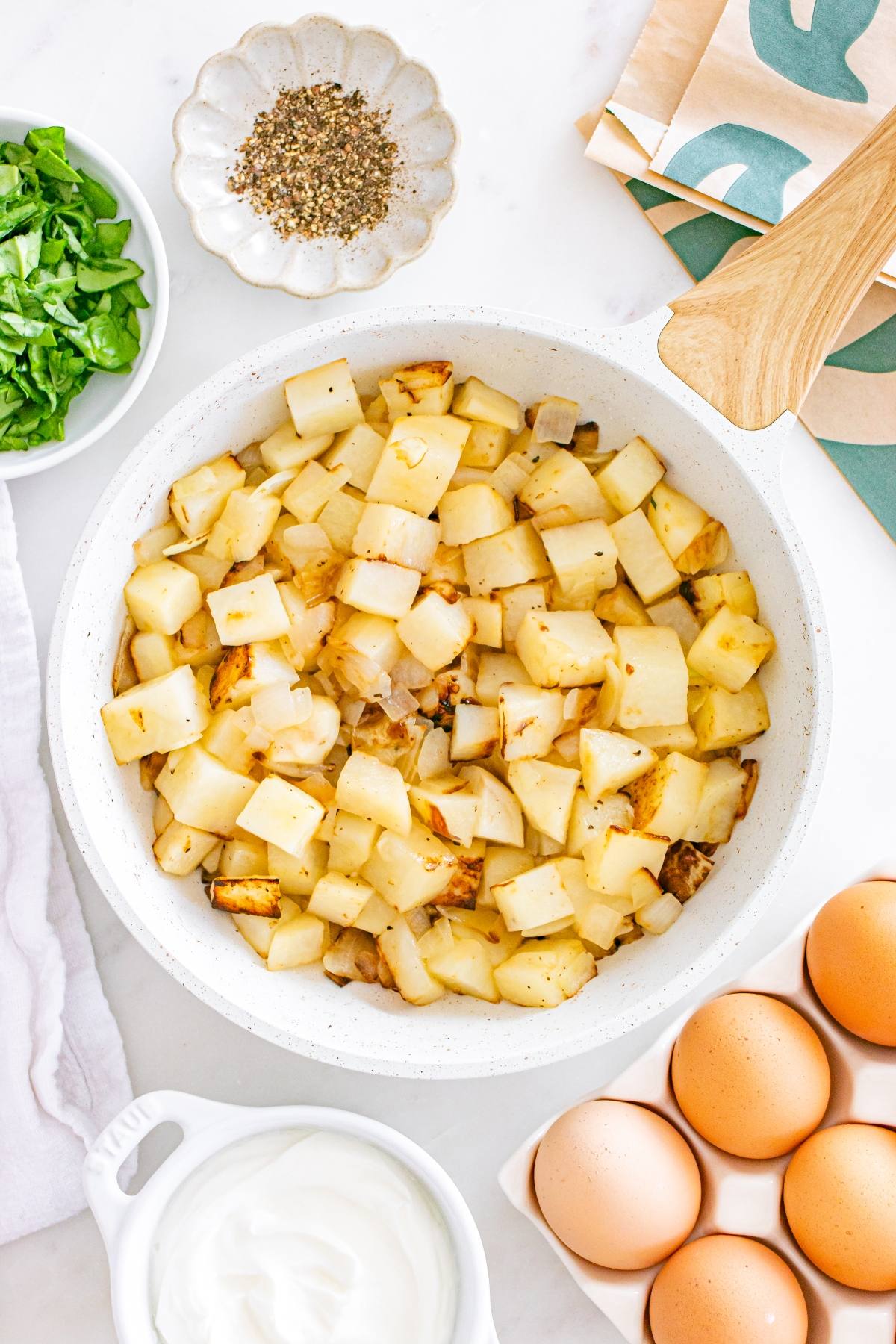 Overhead view of diced potatoes in a skillet, with eggs, spinach, seasoning, and yogurt on a white surface.