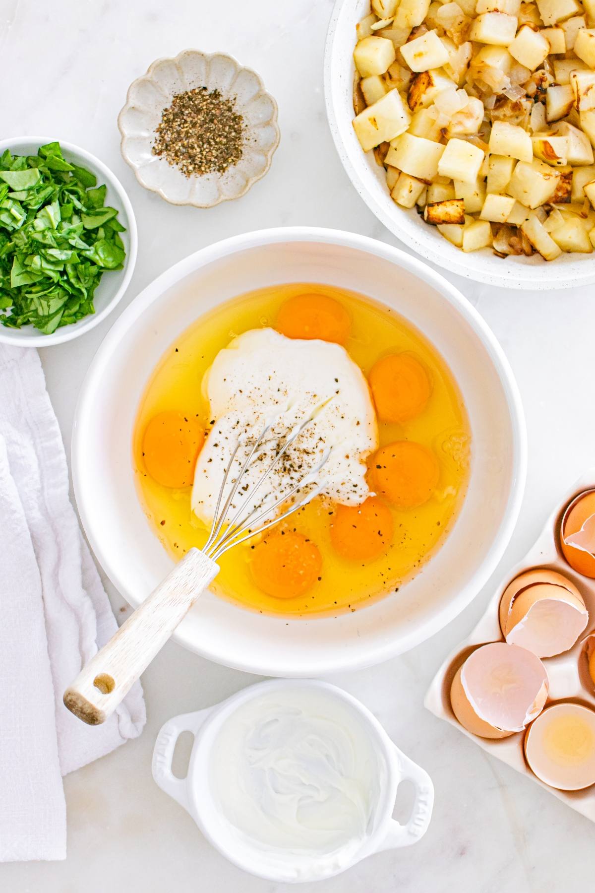 Bowl with eggs, cream, and pepper being whisked, surrounded by potatoes, spinach, eggshells, and seasonings.