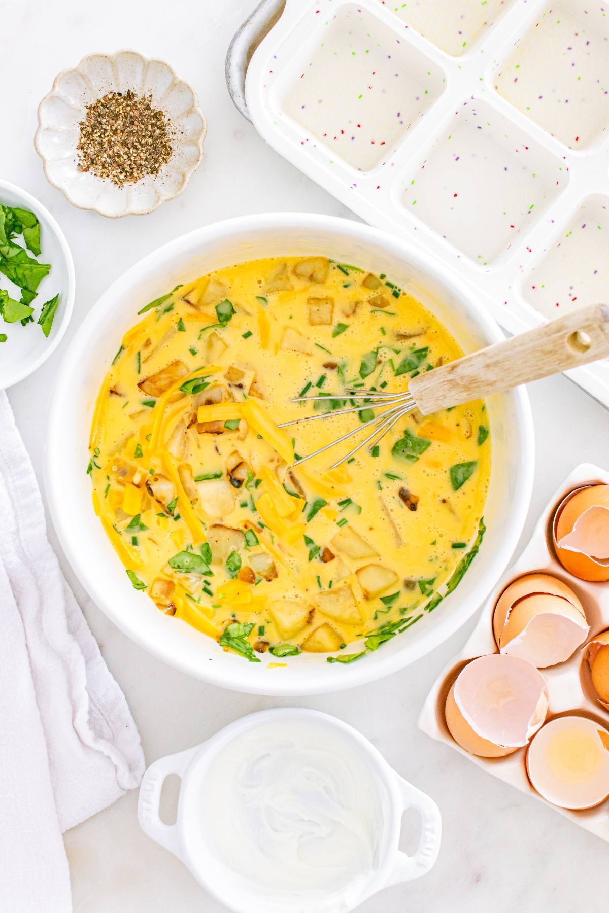 A bowl of whisked eggs with vegetables, herbs, and cheese, surrounded by eggshells and seasoning.