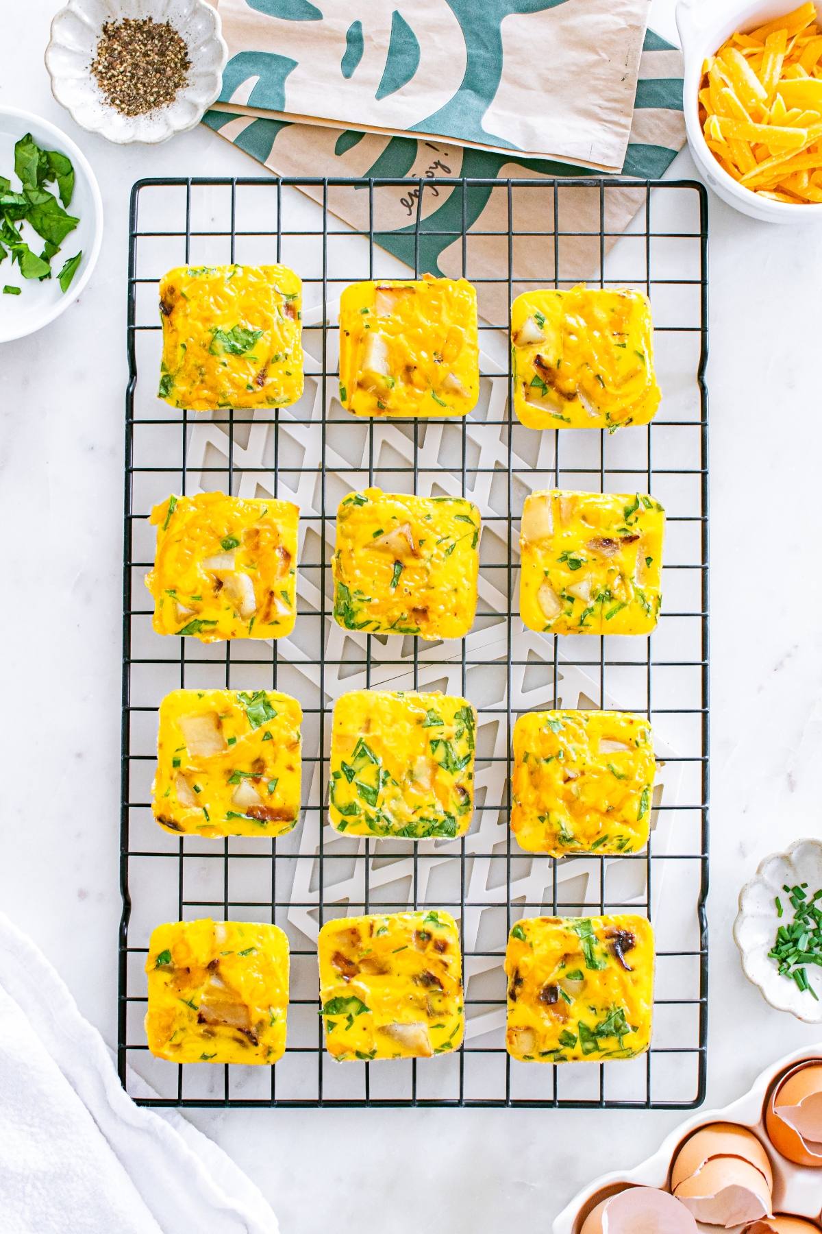 Twelve square egg baked bites with herbs and cheese cooling on a wire rack, surrounded by ingredients.