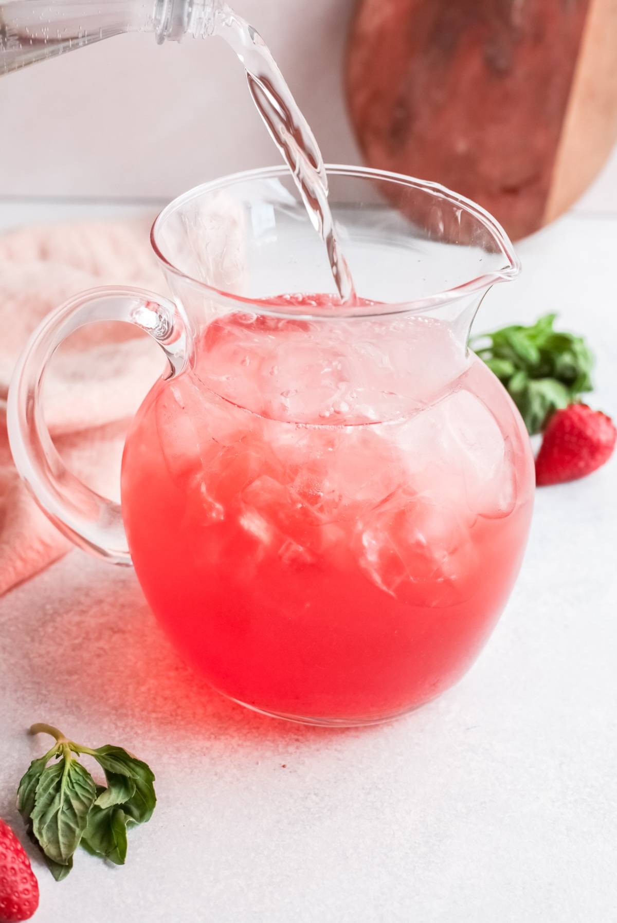 A glass pitcher of pink lemonade with ice being poured, surrounded by strawberries and basil leaves—a refreshing variation on a classic gin rickey made with strawberry basil simple syrup.