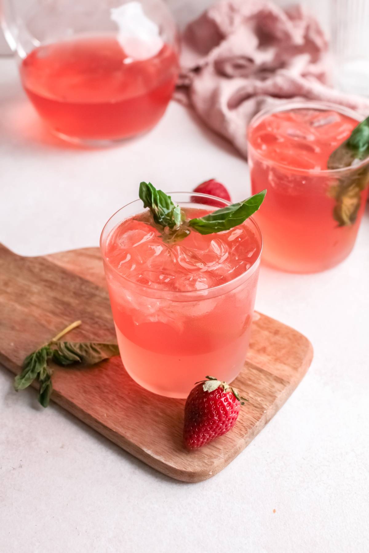 A glass of pink iced drink with strawberry basil simple syrup and a strawberry on a wooden board, another glass and pitcher behind—perfect for serving as a refreshing summer cocktail.