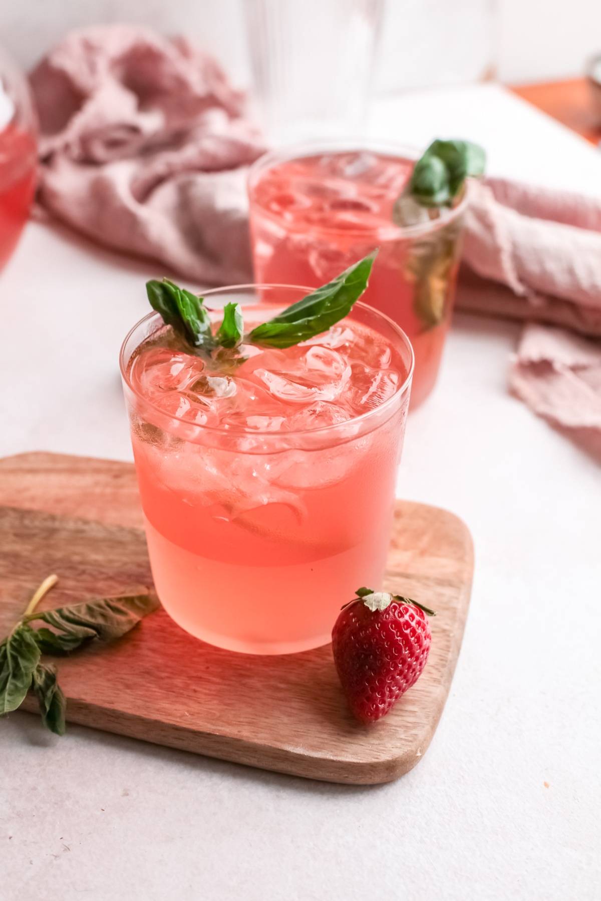Two glasses of pink iced drinks with basil leaves sit on a wooden board, featuring a strawberry and basil nearby—a refreshing summer cocktail made with strawberry basil simple syrup.
