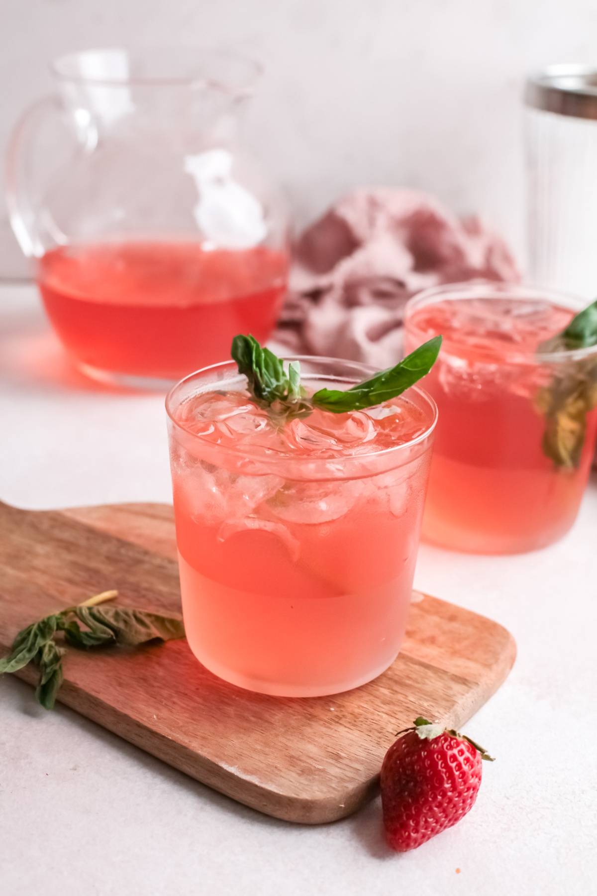 Iced strawberry drink garnished with basil in glasses, with a pitcher and fresh strawberry on the side—perfect as a refreshing summer cocktail or a delightful Strawberry gin rickey.