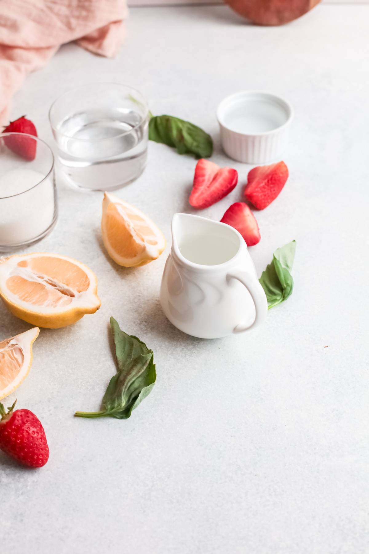 White creamer, lemon wedges, basil leaves, strawberries, water, and sugar on a light surface—perfect for crafting a cocktail with gin or a refreshing variation on a classic gin rickey using strawberry basil simple syrup.