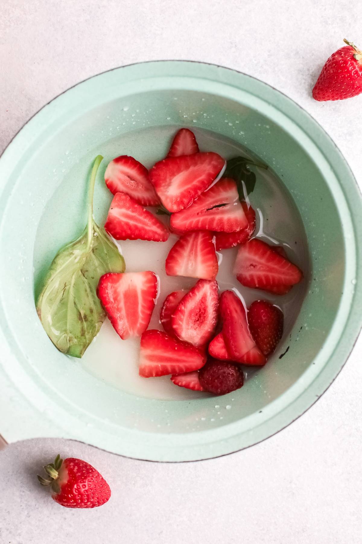 Sliced strawberries, a basil leaf, and liquid in a light green bowl on a white surface—perfect ingredients for a refreshing summer cocktail or strawberry basil simple syrup.