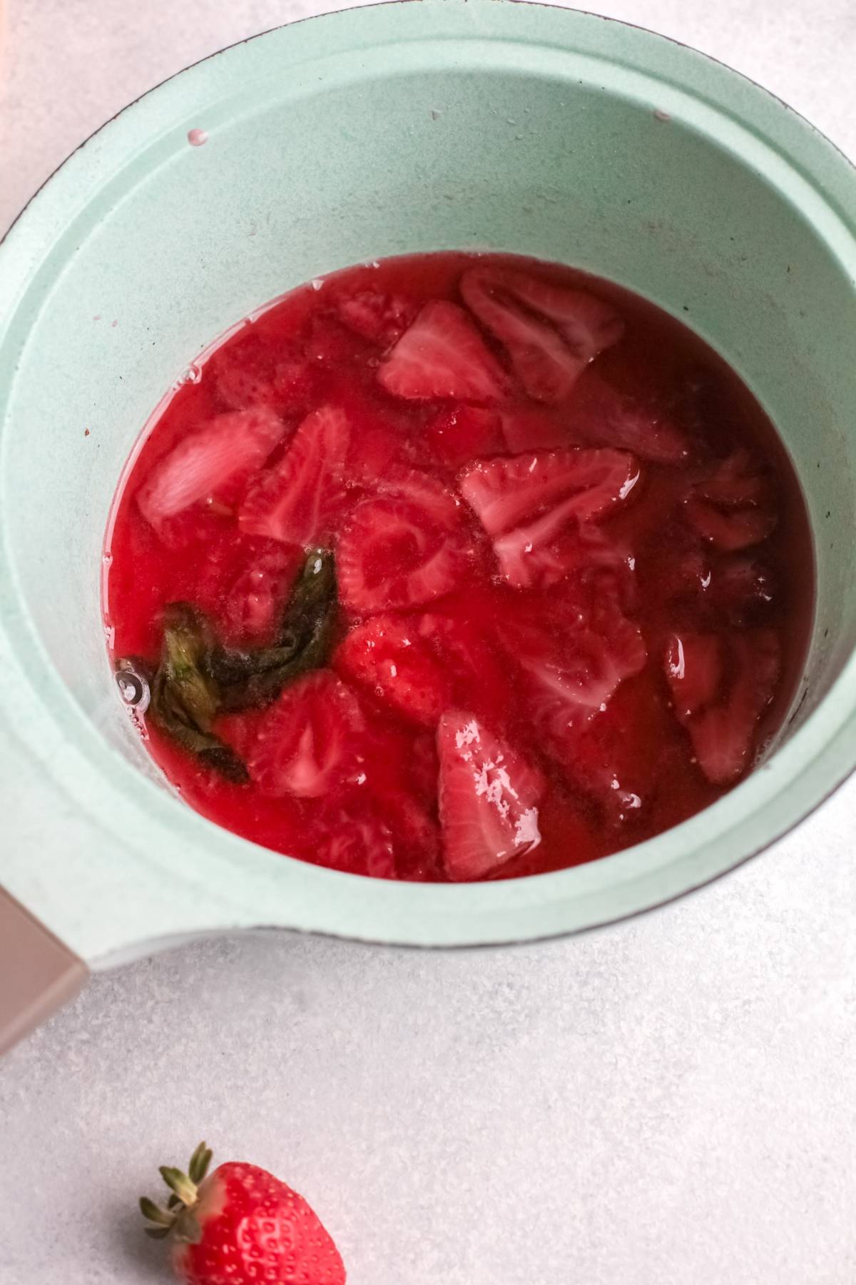 Strawberry compote with sliced strawberries and mint leaves simmering in a light green saucepan—a perfect topping for a refreshing summer cocktail.