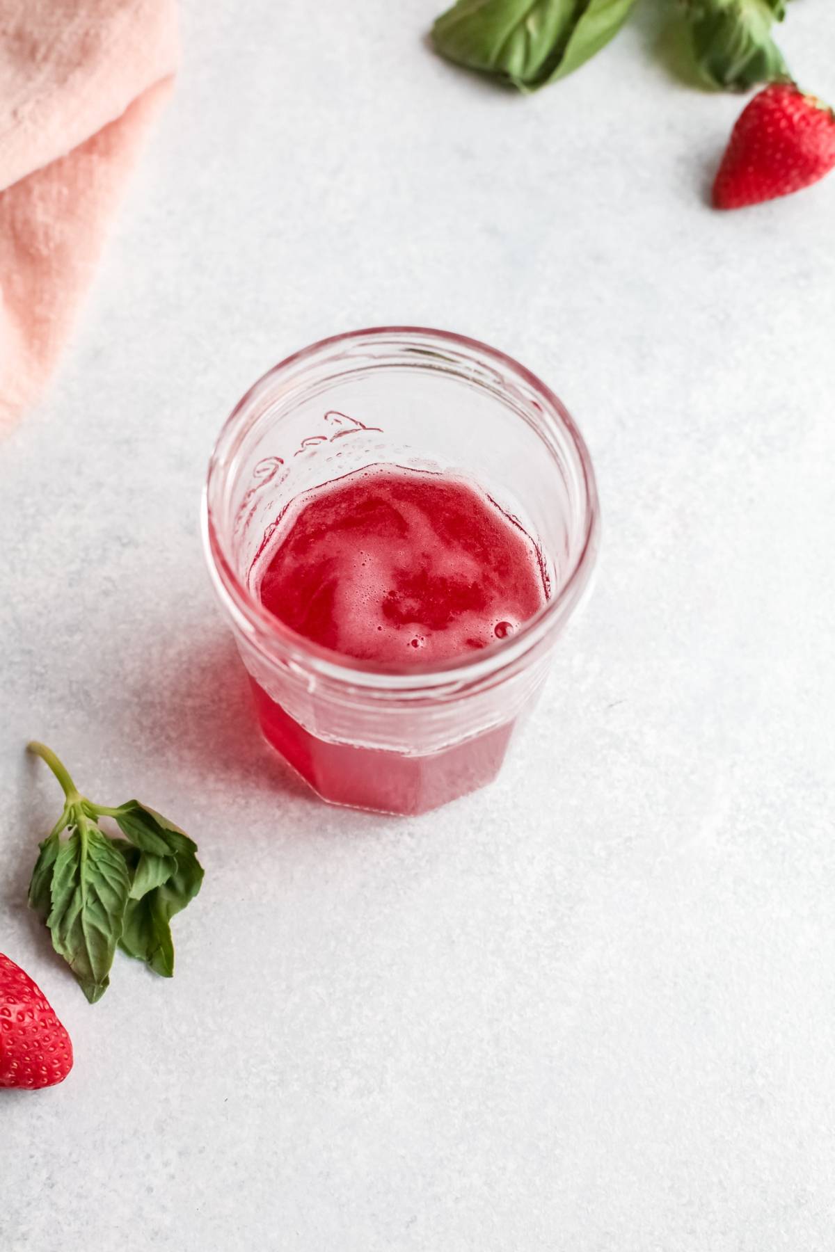 A glass jar with pink juice on a light surface, surrounded by strawberries and basil leaves—a refreshing summer cocktail and a vibrant variation on a classic gin rickey.
